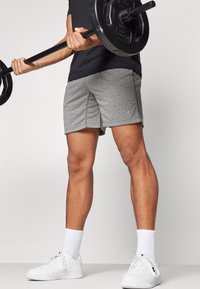 Grau gemusterte Shorts mit strukturierter Oberfläche, kombiniert mit einem schwarzen Hemd und weißen Sneakern. Die Figur hält eine Langhantel.