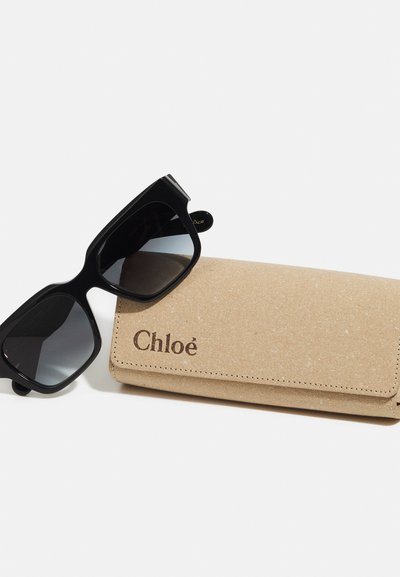 Black sunglasses with a geometric frame and gradient lenses, resting on a tan, textured case embossed with "Chloé."