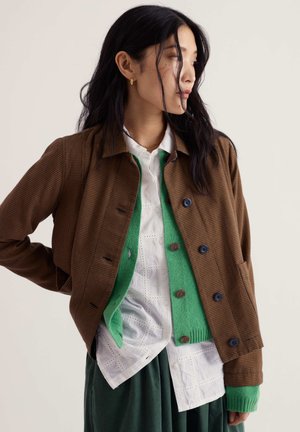 Donna con lunghi capelli neri indossa una giacca marrone a quadri, un cardigan verde, una camicia bianca e una gonna verde, guardando di lato.