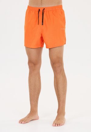 Mand iført lyse orange badeshorts med sorte snore, stående barfodet på hvid baggrund.