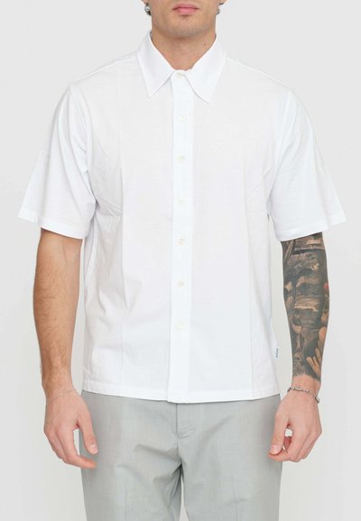 Camicia bianca a maniche corte con colletto a punta, chiusura con bottoni sul davanti e texture morbida, abbinata a pantaloni grigi.