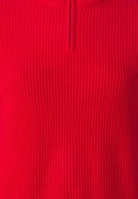 Pull rouge côtelé avec un petit col demi-zip. Le tissu semble texturé avec des lignes verticales et une finition lisse.
