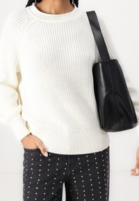 Pull blanc tricoté avec un col rond et des poignets côtelés, associé à un jean en denim noir orné de clous argentés, et un sac à main en cuir noir.