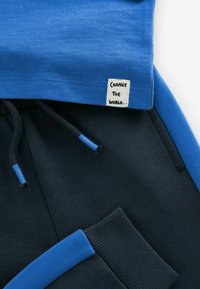 Blå bomulds sweatshirt med ribbet halsudskæring, parret med mørkeblå bomulds joggers med blå accenter og et mærke, der står "FORANDR VERDEN."