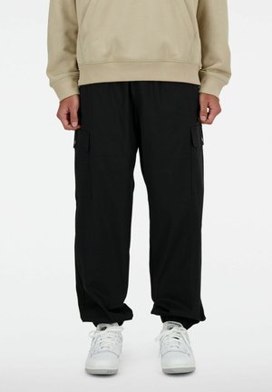 Person iført sorte cargobukser, beige hættetrøje og hvide New Balance sneakers, stående mod en hvid baggrund.