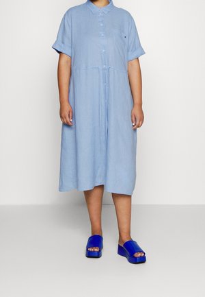 Person trägt ein hellblaues, kurzes, knöpfbares Midikleid mit aufgekrempelten Ärmeln und leuchtend blaue, offene Plateausandalen.