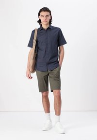 Kurzärmliges, knopfbares Hemd in Marineblau mit dezentem Muster, olivgrüne Shorts, weiße Sneakers und eine beigefarbene Umhängetasche. Einfaches, lässiges Outfit.