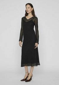 Melna mežģīņu kleita ar garām piedurknēm un V-veida kakla izgriezumu, ar izplestu svārku un mežģīņu apdari ap maliņu. Nēsāta ar norādīta pirksta augstiem papēžiem.