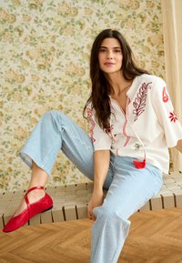 Mujer con cabello largo y oscuro, vestida con blusa blanca con bordados rojos y pantalones de rayas azules, sentada en un banco tejido frente a un papel tapiz floral.