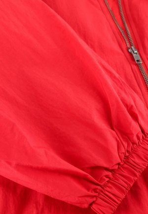 Rode jas met mouw en elastische manchetten, zichtbare zilveren rits op lichtgewicht stof.