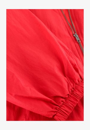 Rode jas met mouw en elastische manchetten, zichtbare zilveren rits op lichtgewicht stof.