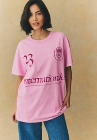 Rozā kokvilnas t-krekls ar apaļu kakla izgriezumu, rotāts ar košu bordo grafisku tekstu un logotipu. Brīvs piegrieziens, apvienots ar augstās jostasvietas džinsiem.