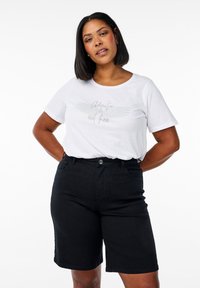 Camiseta blanca con un diseño impreso que dice "La aventura está ahí fuera", combinada con pantalones cortos de denim negro de tiro alto. Textura suave, ajuste casual.