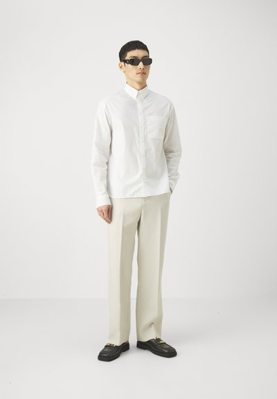 White long-sleeve button-up shirt with a pocket, paired with light beige trousers and black shoes featuring gold accents. Black sunglasses complete the look.
