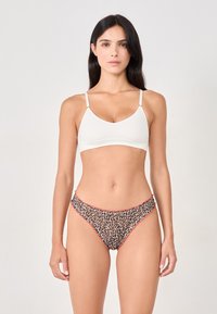 Woman with long dark hair wears white bralette and leopard-print panties with red trim, standing against plain light background.