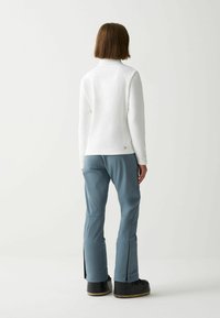 Maglia bianca a maniche lunghe e pantaloni da sci blu chiaro con dettagli zip e vestibilità comoda. Stivali da neve neri completano l'outfit.