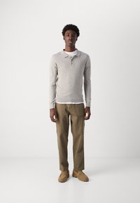 Grå langærmet polo med tre knapper, parret med lyse khaki bukser og beige slip-on sko, stående mod en neutral baggrund.
