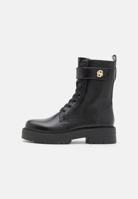Botas de cuero negro hasta el tobillo con puntera redonda, cierre de cordones en la parte frontal, cuello acolchado, acentos en hardware dorado y suela gruesa de goma negra.