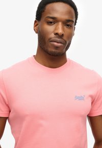 Roze katoenen t-shirt met een ronde hals, korte mouwen en een klein blauw geborduurd logo op de linker borst. Glad textuur.