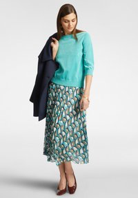 Maglione turchese con texture, gonna plissettata a stampa nei toni del blu, verde e marrone, tacchi marroni, giacca scura appoggiata su un braccio.