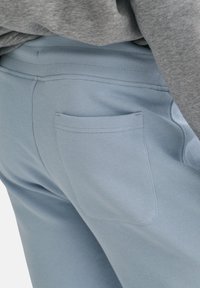 Pantalon de survêtement bleu clair avec une poche arrière, doté d'une texture douce et d'une taille élastique. Un tissu gris est visible au-dessus du pantalon.