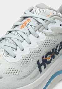 Sapatilha atlética cinza claro com parte superior em rede, atacadores tonais e um proeminente logo HOKA em azul marinho escuro com um destaque laranja. Apresenta uma sola branca espessa.