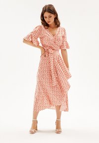 Rode en witte polka dot wikkeljurk met korte gerimpelde mouwen en een asymmetrische zoom. Gemaakt van lichte stof, strakker getrokken in de taille.