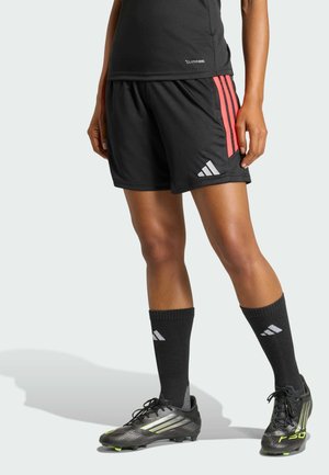 Person trägt schwarze Sportshorts mit pinken Streifen, schwarze Adidas-Socken und schwarze Fußballschuhe und steht vor hellem Hintergrund.