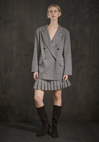Grijze geruite dubbelbreasted blazer met ingezette revers, gelaagd over een geplooide minirok. Het model draagt kniehoge bruine suède laarzen.