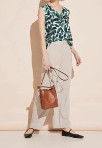 Woman wearing sleeveless green leaf print top with beige wide-leg pants, holding a brown leather handbag, and wearing black flats.