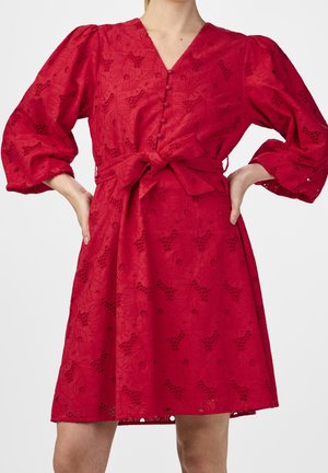 Kobieta w czerwonej sukience do kolan z bufkami na rękawach, guzikami z przodu, dekoltem w serek, kwiatowym wzorem z ażurowym haftem oraz kokardą na talii.