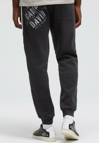 Dunkelgraue Sweatpants mit gerippten Bündchen, seitlichen Streifen und weißem Grafiktext. Getragen mit schwarz-weißen Sneakers mit Logo-Akzenten.