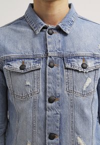 Blekt blå denimjacka med en klassisk krage, fem metallknappar, två bröstfickor och slitna detaljer på tyget.