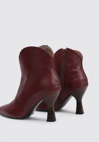 Botas de tobillo de cuero burdeos con acabado texturizado, punta afilada y un tacón de madera de 5 cm. Cuenta con un cierre de cremallera en la parte posterior.