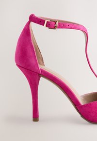 Rózsaszín velúr magas sarkú cipő bokapánttal és arany csattal. Keskeny orrú és vékony tűsarkú. Belső része sima bélésű.