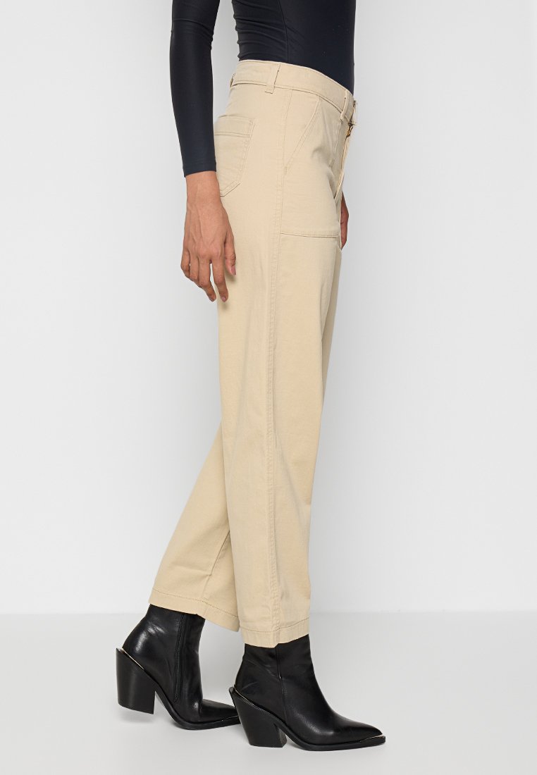 Pantalones de pierna ancha beige hechos de una mezcla de algodón, con bolsillos laterales y cintura alta, combinados con botas de tobillo negras de tacón cuadrado.