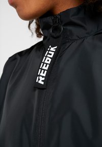 Chaqueta negra con cremallera y cuello alto, que presenta una etiqueta con el logo de Reebok y un material liso y ligero. Vista en primer plano que muestra los detalles de la cremallera.