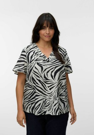 Frau mit langen dunklen Haaren, die eine kurzärmelige schwarz-weiße Zebramuster-V-Ausschnitt-Bluse trägt und vor einem schlichten hellen Hintergrund steht.