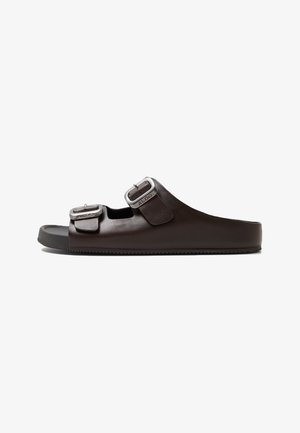 Brown slide sandal with dual adjustable metallic buckles, made from smooth synthetic material, featuring a textured rubber sole.