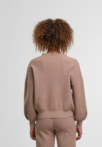 Pull en maille à manches longues de couleur marron clair, avec un motif texturé, des épaules tombantes et un ourlet côtelé. La vue de dos montre une coupe décontractée.