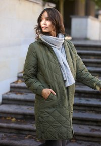 Cappotto imbottito verde con cappuccio, dotato di polsini a coste, tasche laterali e una sciarpa grigia chiara indossata sotto. Il modello si trova vicino a delle scale.