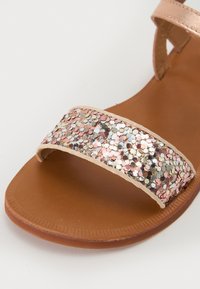 Flat sandal med en brun såle og et bredt glitterbånd som har rosa, sølv og mintfargede heksagonale pauser. Stroppene er tan og glatte.