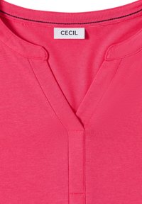 T-shirt en coton rose à col en V avec une étiquette portant l'inscription "CECIL." Présente une texture lisse et des accents cousus autour de l'encolure et de la patte de boutonnage.