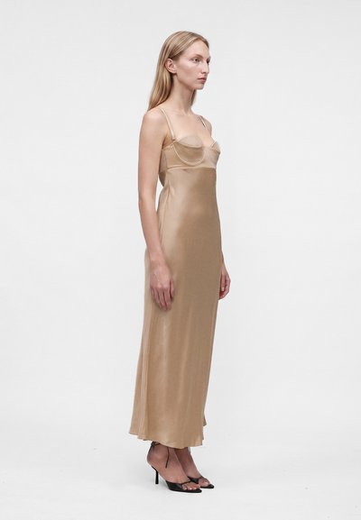 Femme en longue robe nuisette en satin beige à fines bretelles et sandales noires à talons hauts, debout de profil sur fond uni.