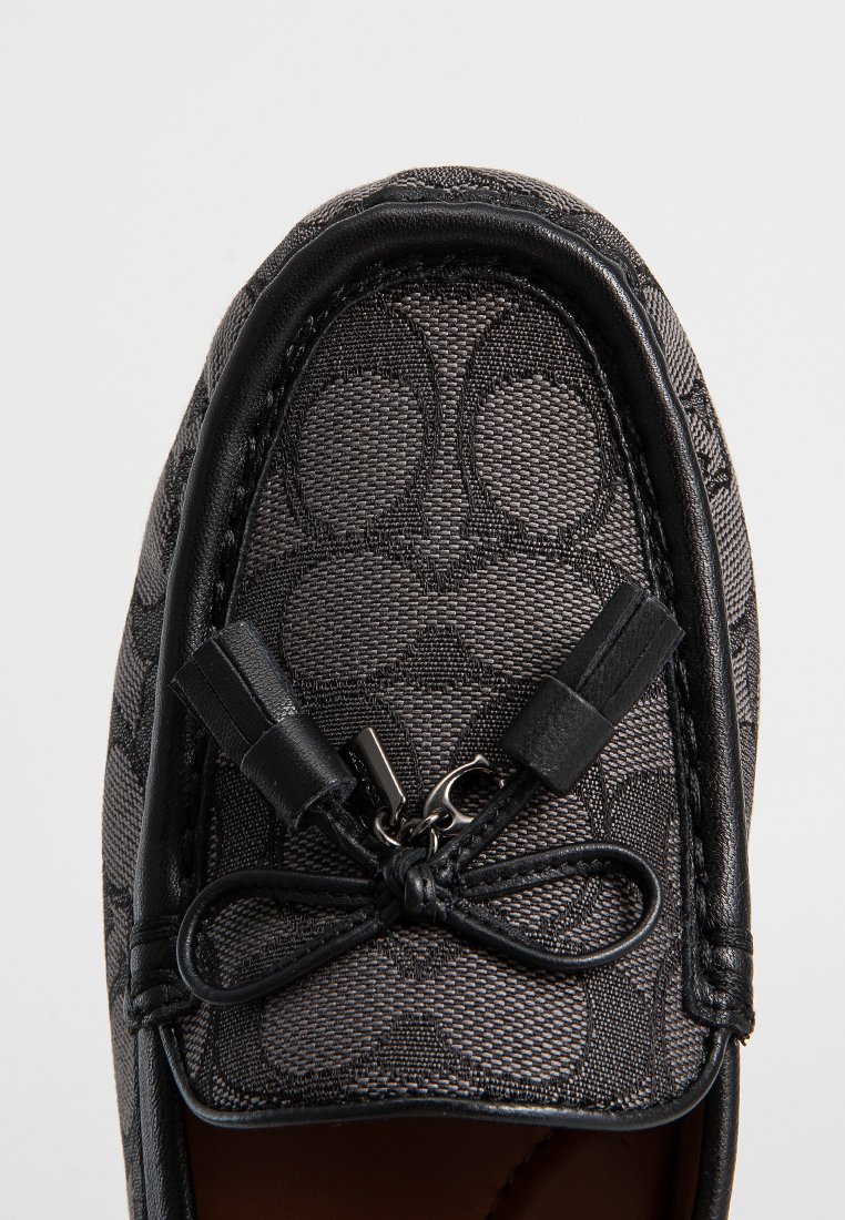 Zwarte textiele loafers met een subtiel geëmbossed patroon, leren accenten en een decoratieve strik met metalen hardware. Gladde textuur en klassiek ontwerp.