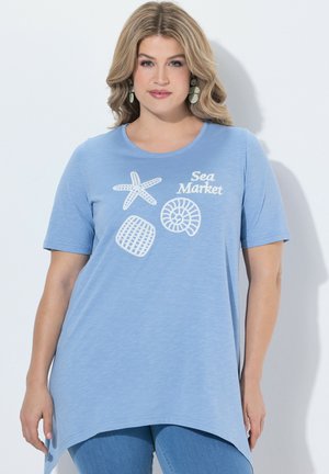 Donna che indossa una t-shirt asimmetrica azzurro chiaro con stampe bianche a tema marino e scritta "Sea Market", abbinata a jeans blu.