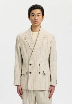 Giovane uomo che indossa una giacca doppiopetto beige a righe sottili e pantaloni abbinati, in piedi davanti a uno sfondo chiaro e uniforme.