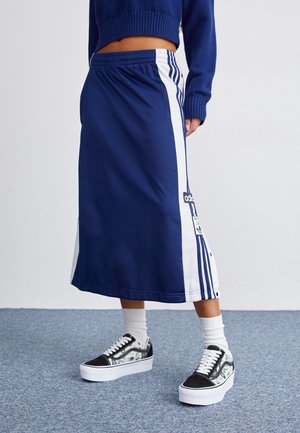 Femme portant une jupe Adidas bleu marine et blanche avec des bandes latérales et un patch décoratif, des chaussettes blanches, et des baskets à plateforme noir et blanc.