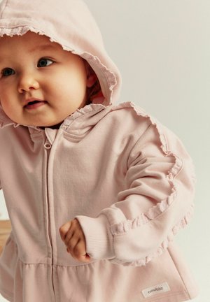 Jasnoróżowa bluza z kapturem z falbaną i teksturowaną tkaniną. Posiada przedni zamek błyskawiczny i metkę na dolnym brzegu.