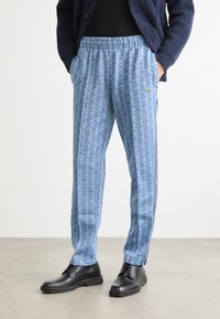 Blå mønstrede joggebukser med geometrisk design, elastisk midje, sidelommer og kontraststripe. Svart sko på.